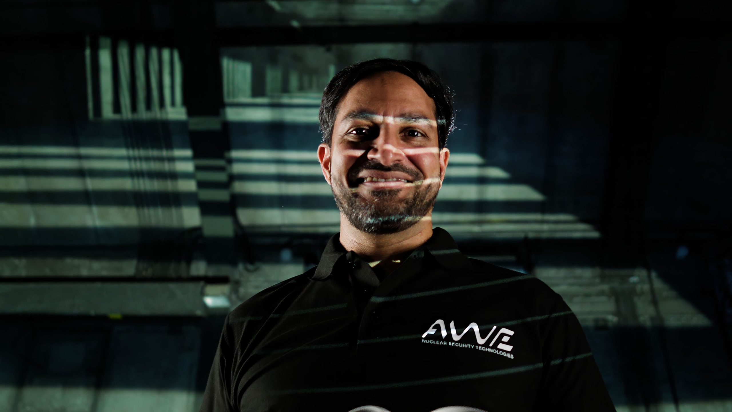 The image depicts a person standing in a dimly lit setting, with shadows and light patterns creating a moody background. The individual's face is intentionally blurred for privacy. They are wearing a black polo shirt featuring the logo "AWE" and the text "AWE Security Technologies"