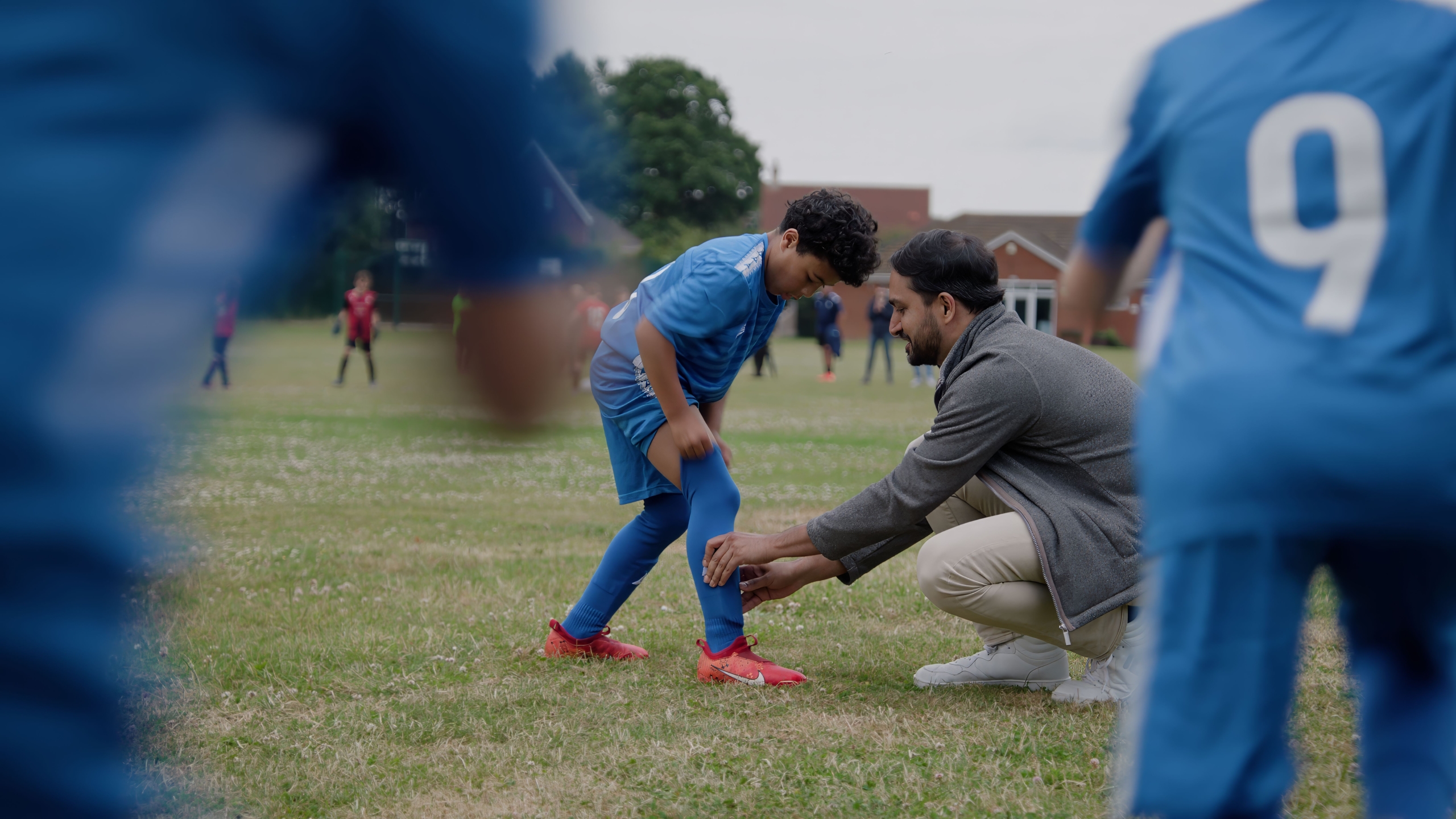 Man in a gray jacket and beige pants kneeling on grass, assisting a young football player in a blue uniform with red cleats in adjusting their shin guard; other players and buildings visible in the background