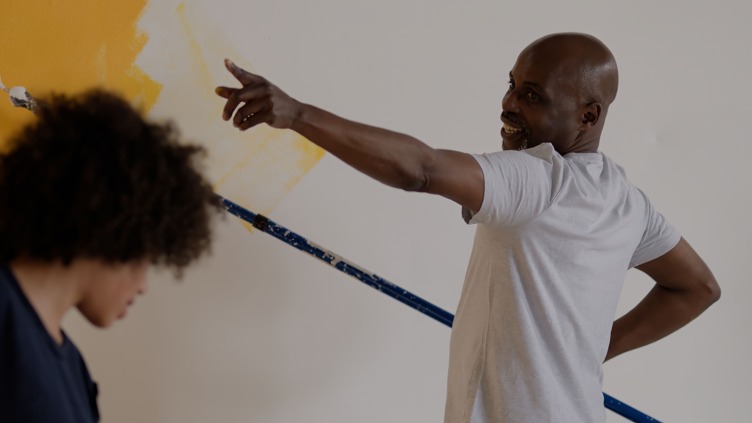Two people painting a wall; one holds a roller with yellow paint and points toward the wall, while the other looks down. The wall is partially painted yellow.