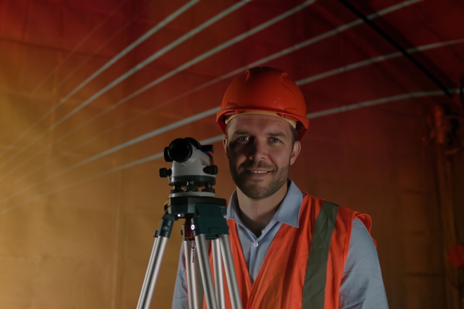 A person in an orange hard hat and safety vest smiles at the camera and uses a surveying instrument on a tripod in a dimly lit industrial setting