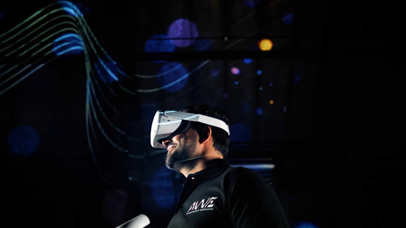 Person wearing a virtual reality headset, dressed in a black 'AWE' shirt, standing against a dark background with colourful light patterns and lines.