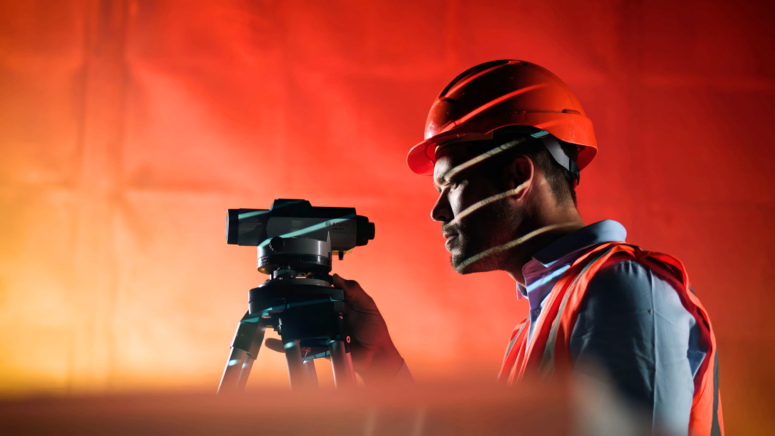 Close-up of a person in a red hard hat looking through a surveying instrument, with an orange background and light streaks casting shadows on the face