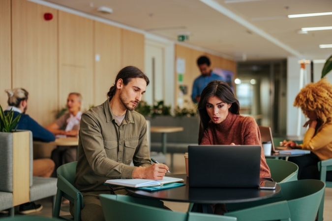 Two young coworkers are on a working breakfast in the company cafeteria, working on a laptop and catching notes in a notebook.