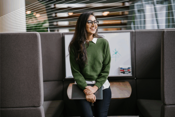 Portrait of a young woman student smiling as she leans in a discussion booth in her campus. She is wearing a green jumper.
