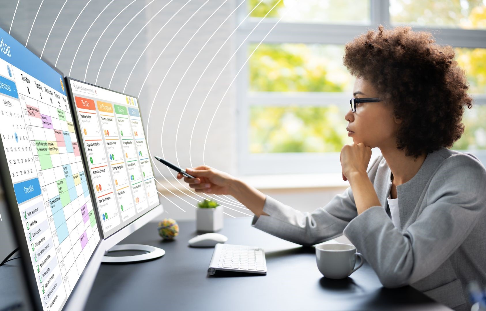 Person working at a desk with dual monitors, reviewing a weekly schedule and project tasks. The workspace includes a coffee cup, keyboard, and small plant, with a window showing greenery outside
