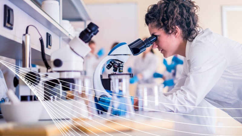 Feature image rainee in a white lab coat examines samples under a microscope in a research lab filled with scientific instruments and glassware, illustrating hands-on training in nuclear materials analysis.