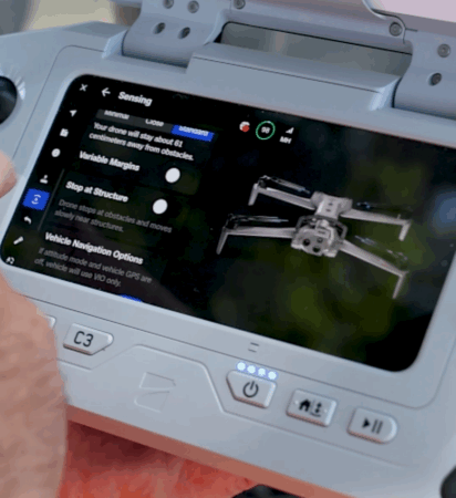A close-up of a drone controller screen showing sensing settings such as Variable Margins, Stop at Structure, and Vehicle Navigation Options. The display includes an image of a drone on the right side. The controller features buttons like the power button and C3 button, with a person’s hand partially visible holding the device.