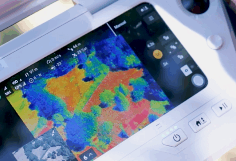 A close-up of a device screen showing a heat map with red and yellow areas indicating higher temperatures and blue and green for cooler areas. The display includes flight data such as altitude, speed, and distance, along with a small inset aerial view of the surveyed area.