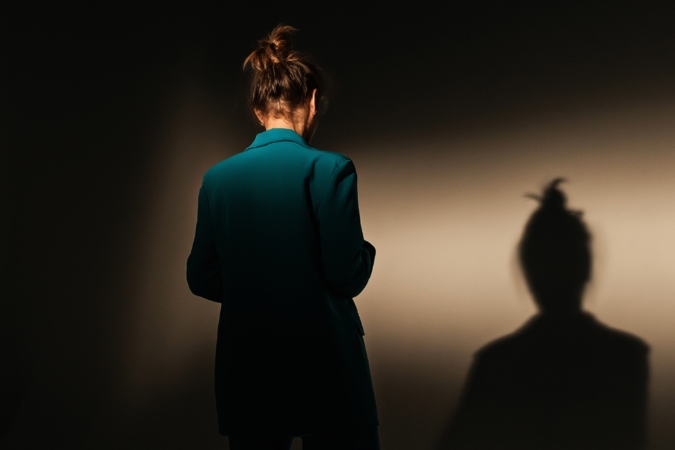 Person standing with their back to the camera, wearing a dark green blazer and hair in a bun, casting a prominent shadow on a plain dark wall."**

Would you like a version tailored for accessibility, SEO, or social media use?