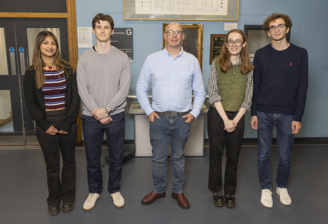 Five people standing indoors in front of a blue wall with framed documents and plaques. They are dressed casually and facing the camera, standing on a grey floor near a door with glass panels.