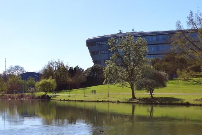 Feature image Curved glass building on a grassy hill with trees, reflected in a pond under a clear blue sky.