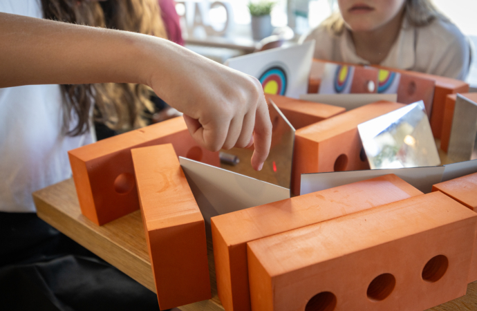 Close-up of a hand interacting with a STEM activity involving orange blocks and angled mirrors on a wooden table. Colorful circular patterns are reflected in the mirrors, suggesting an educational science experiment focused on light and reflection.