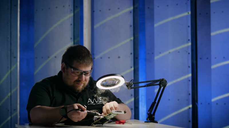 Individual seated at a workstation performing precision soldering on electronic components. A circular task light illuminates the workspace, with AWE branding visible on the dark shirt and a blue-toned industrial backdrop behind.
