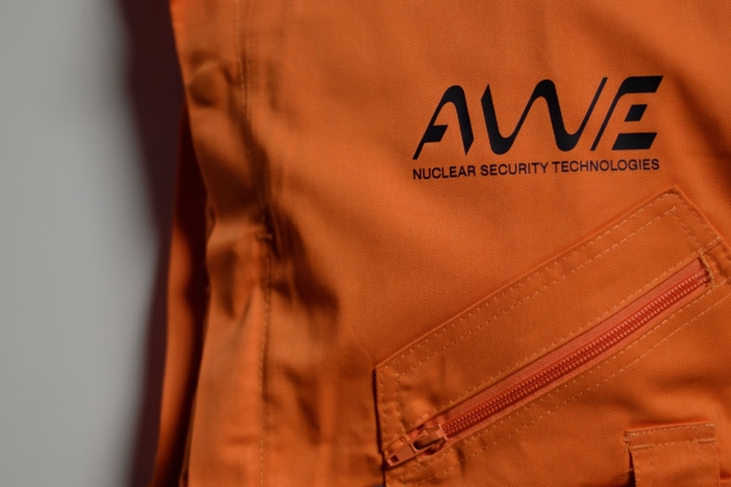 Close-up of an orange protective suit featuring the AWE logo and the words “Nuclear Security Technologies.” A zippered pocket is visible on the front, highlighting the branded workwear used in high-security engineering environments.