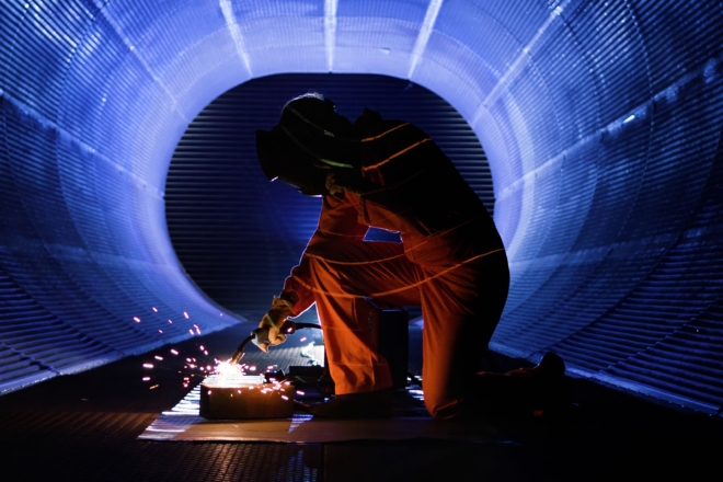A person in protective gear kneeling inside a large cylindrical structure while welding, with bright sparks illuminating the dark interior. The tunnel-like setting is bathed in blue light, creating a dramatic industrial scene.