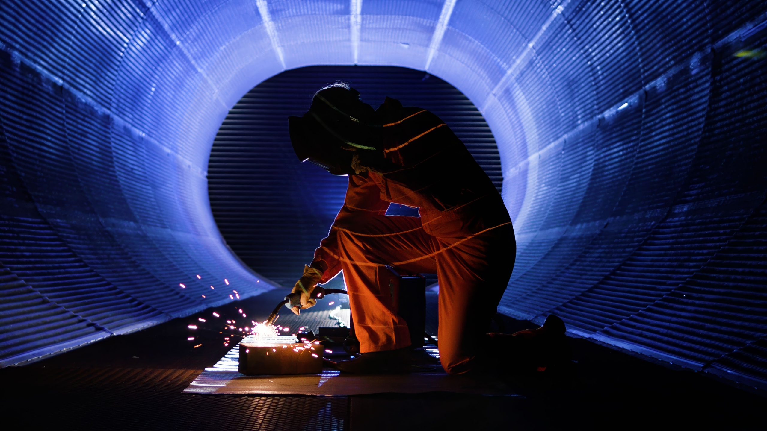 A person in protective gear kneeling inside a large cylindrical structure while welding, with bright sparks illuminating the dark interior. The tunnel-like setting is bathed in blue light, creating a dramatic industrial scene.