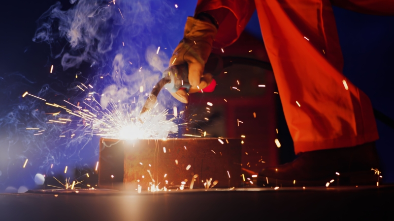 Close-up of welding work with bright sparks and smoke rising from the metal surface. A gloved hand holds the welding torch, and the person is wearing an orange protective suit, emphasizing a high-precision industrial process.