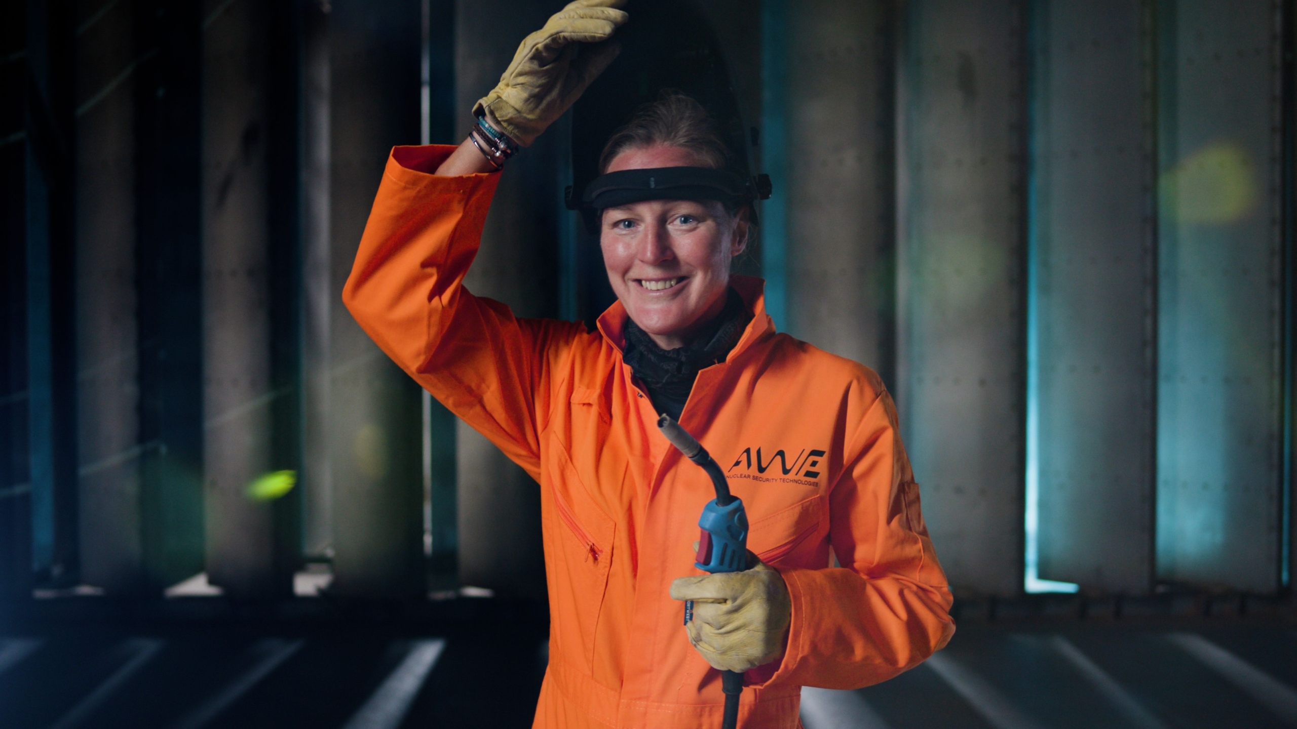 Person wearing an orange protective suit with AWE branding, holding a welding torch in an industrial setting. Vertical metal panels and soft blue lighting form the background, emphasizing a secure engineering environment.
