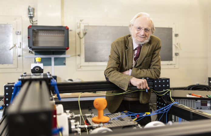 Dr Hugh Goyder in a tweed jacket and tie stands in a laboratory workshop, leaning on a large industrial frame surrounded by cables, machinery, and electronic equipment.