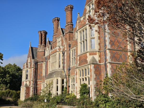 Aldermaston Manor - A large Victorian-style red‑brick manor house with tall decorative chimneys, ornate stone window frames, and multiple gabled roofs, set against a bright blue sky and surrounded by trees and shrubs.