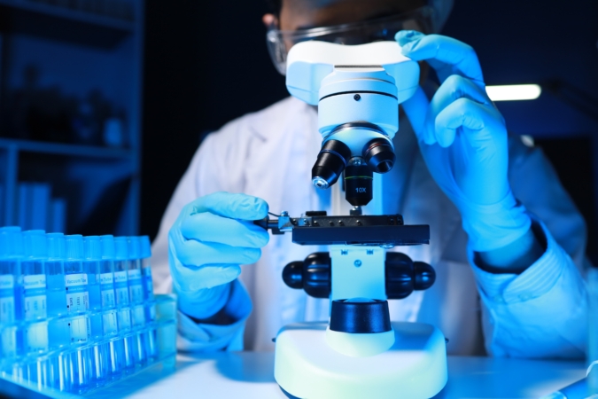 A laboratory setting showing a person wearing protective gloves and eyewear operating a microscope. The individual is adjusting a slide on the microscope stage. Test tubes in a rack are visible beside the microscope, illuminated by blue laboratory lighting.